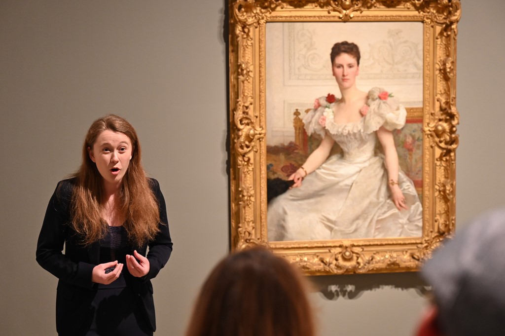 Woman speaking to an audience beside a painting inside an art museum gallery