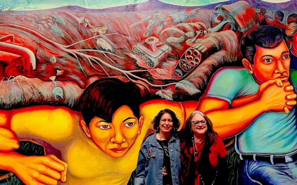 Two women pose in front of a colorful mural